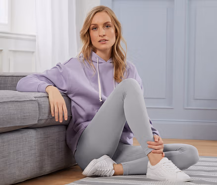 Une femme blonde est assise par terre à côté d'un canapé gris, portant un sweat à capuche violet et un legging côtelé.