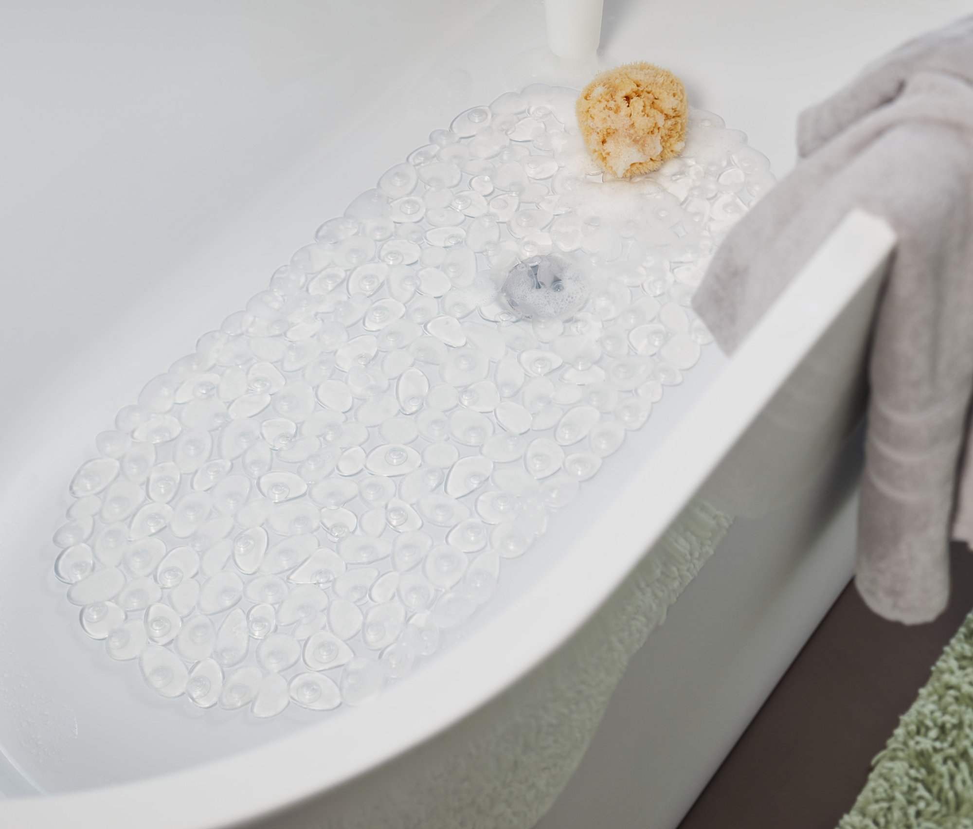 Un tapis de baignoire transparent est posé dans la baignoire. Une serviette grise est suspendue au bord de la baignoire et un tapis vert est posé à côté de la baignoire.