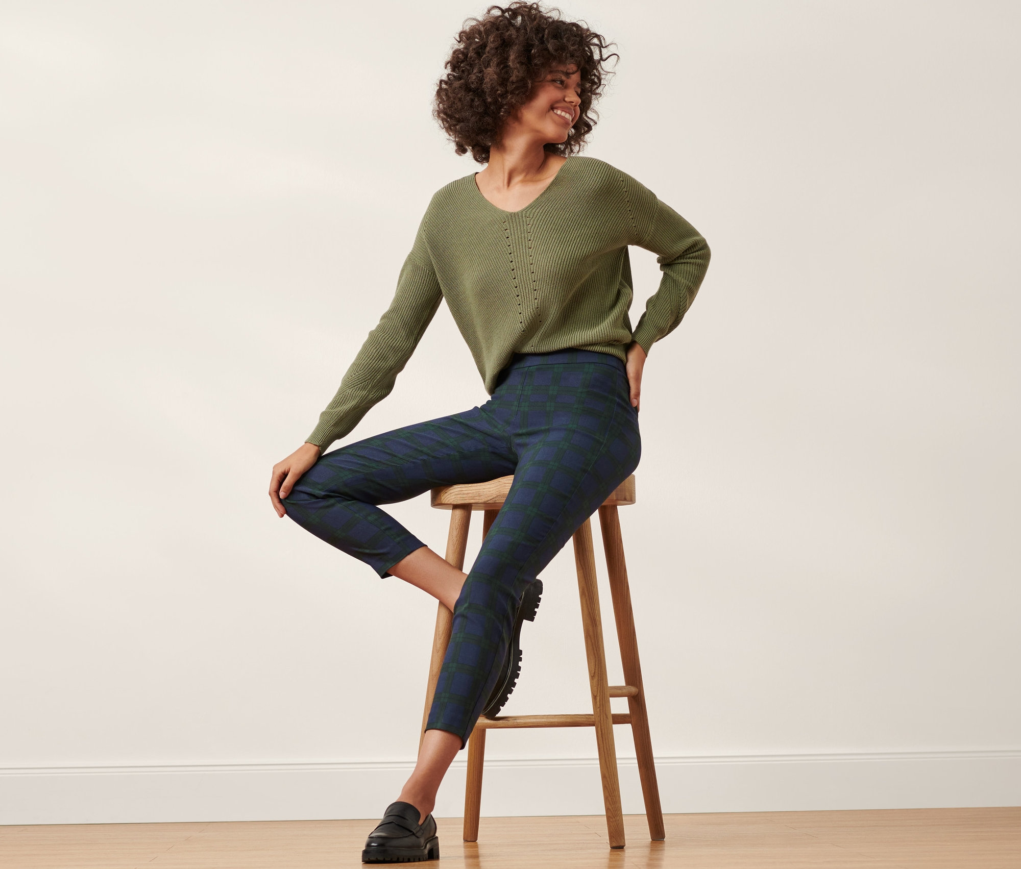 Femme assise sur un tabouret portant un pull vert et un pantalon stretch, à carreaux verts et bleus.
