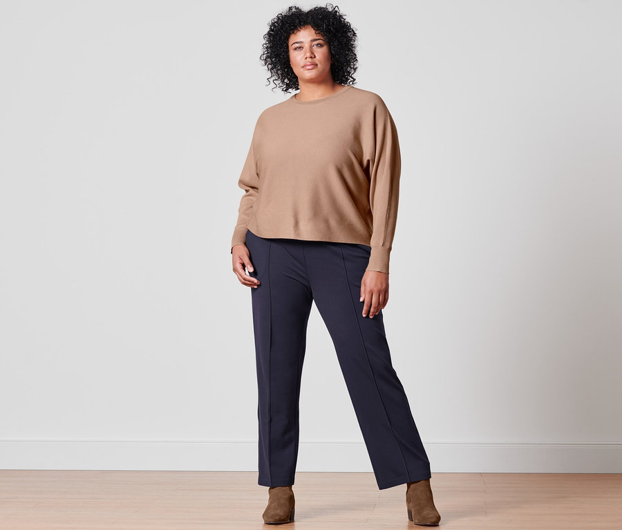 Femme posant en pull en maille fine beige, pantalon en jersey bleu foncé avec nervure et bottines en cuir velours.