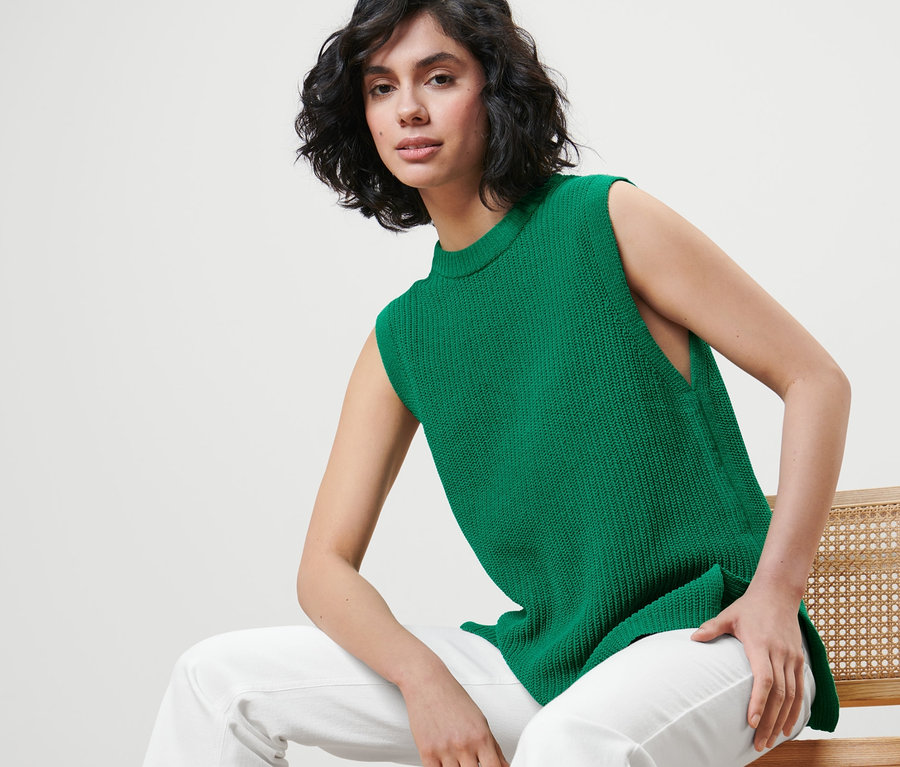 Femme assise sur une chaise portant un pull sans manches en grosse maille vert.