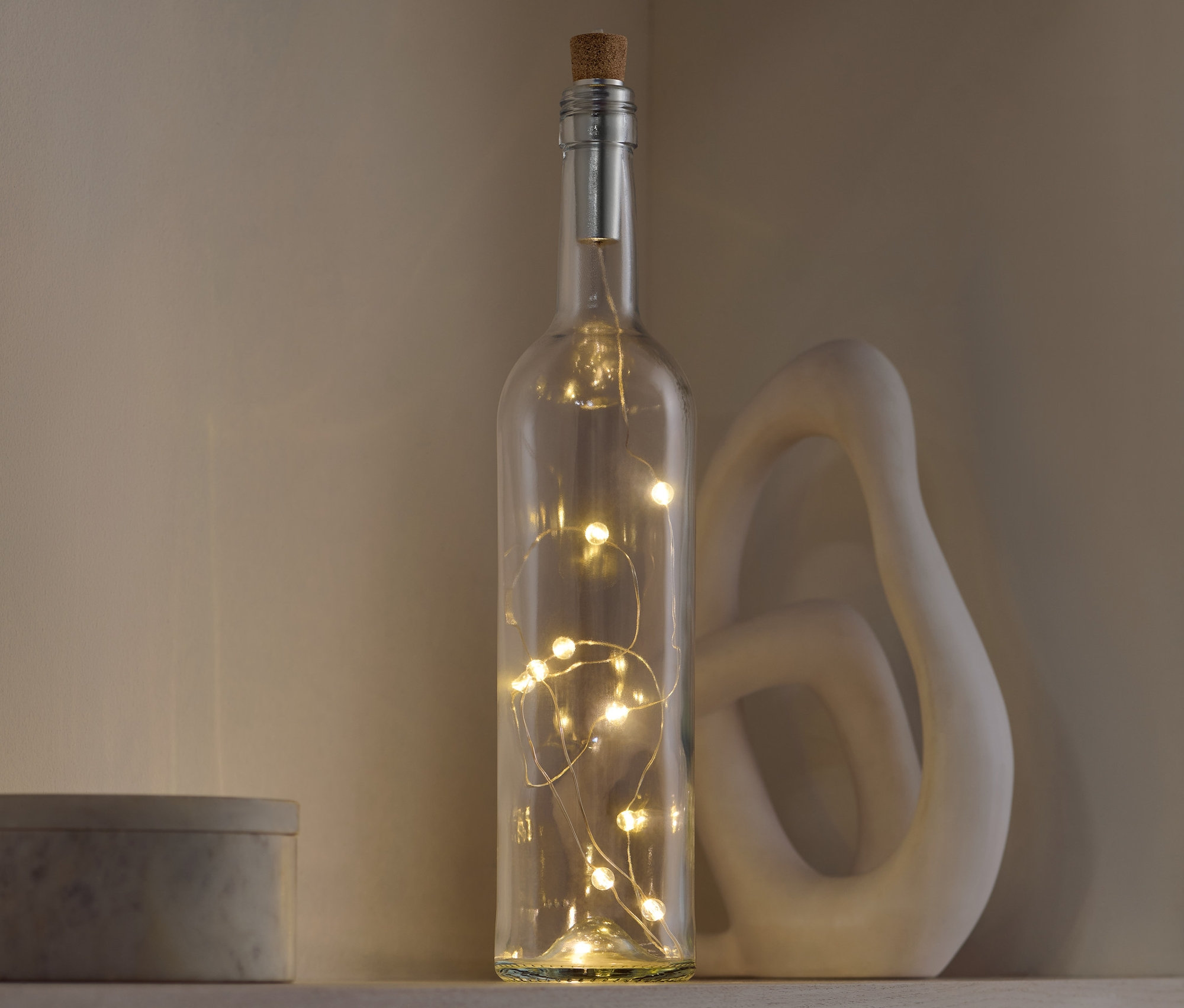Une bouteille décorative en verre avec un bouchon en liège et des lumières à l'intérieur, posée sur une étagère à côté d'une sculpture décorative et d'une boîte grise.