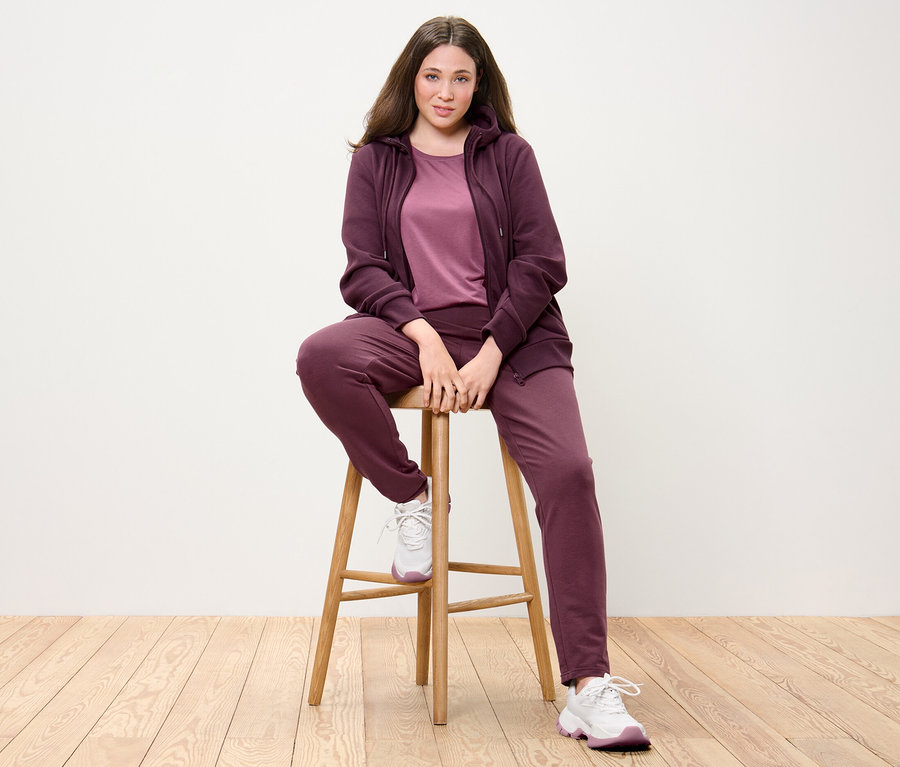 Femme assise sur une chaise portant une jaquette en sweat, un t-shirt long mauve et un pantalon en sweat prune.