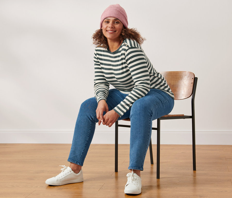 Une femme est assise sur une chaise et porte un t-shirt manches longues à rayures, un jean slim et un bonnet en maille avec de la laine, rose.