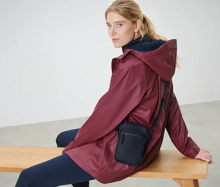 Une femme blonde est assise sur un banc, portant une veste bordeaux et un sac à bandoulière de pluie noir.