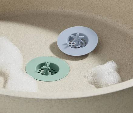 Deux tamis d’évier en silicone sont posés dans l'évier, 2 pièces, un vert et un bleu, entourés de mousse.