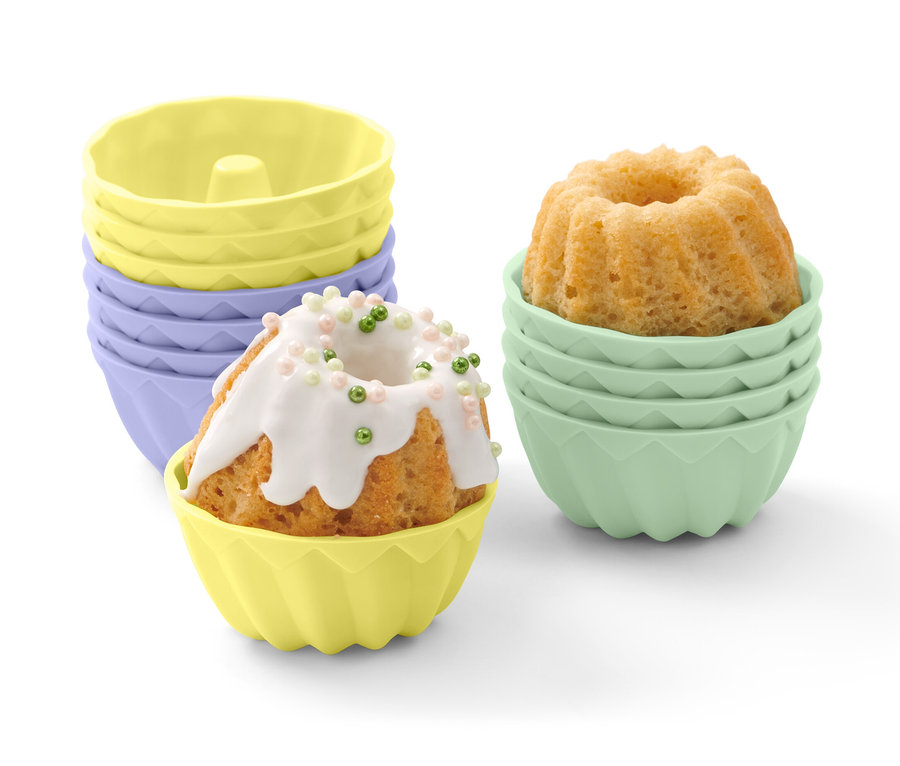 Un mini-kouglof dans un moule jaune, avec un glaçage blanc et des décorations colorées. Autres 12 mini-moules à kouglof en arrière-plan.