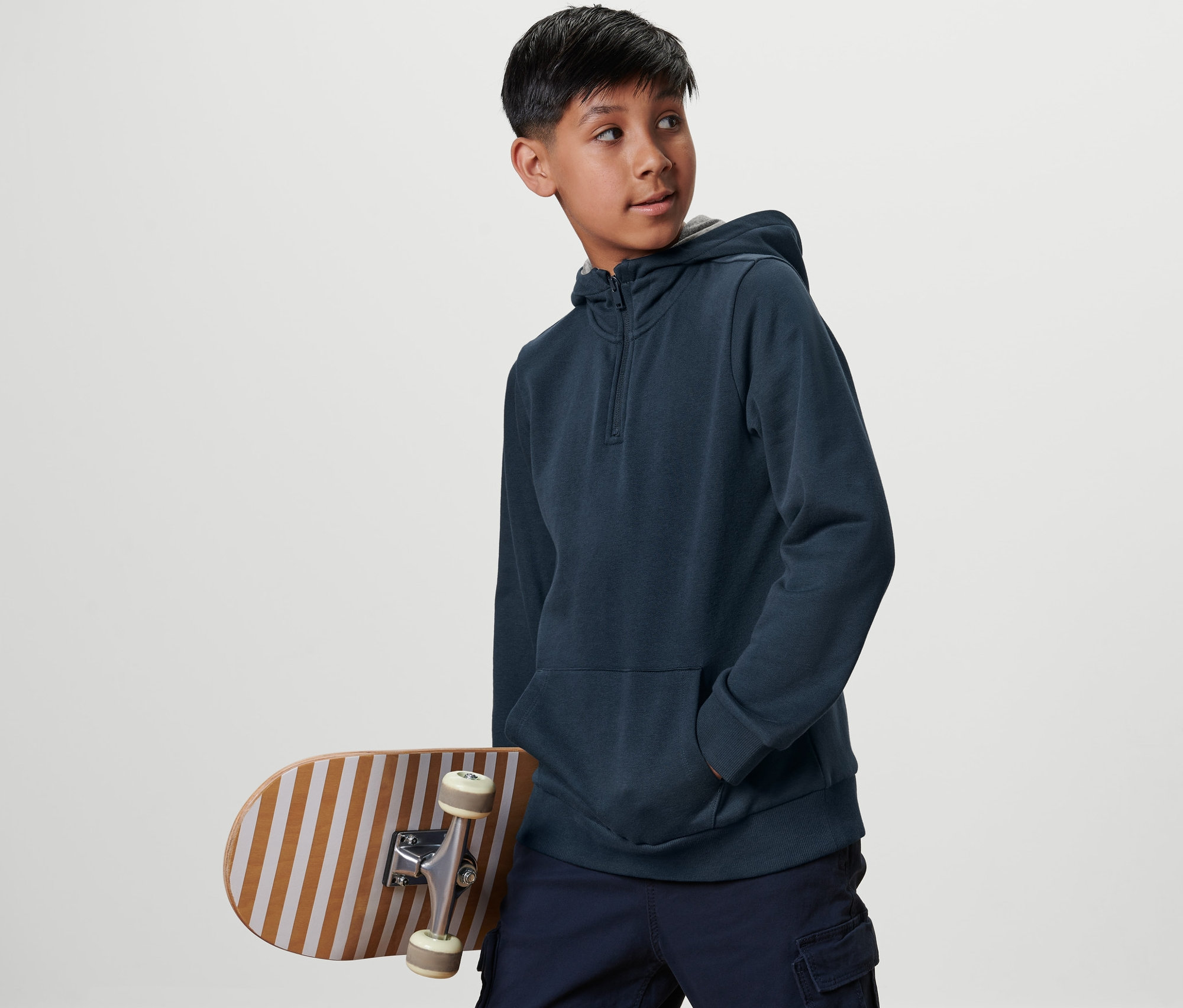 Un garçon avec un skateboard porte un sweat à capuche bleu pour enfant et regarde sur le côté.