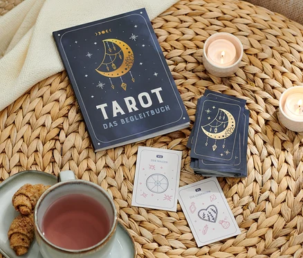 Sur une table en osier se trouvent Tarot : le kit pour débutants avec des cartes et un livre d’accompagnement, des cartes de tarot, deux bougies, une tasse et une assiette avec des croissants.