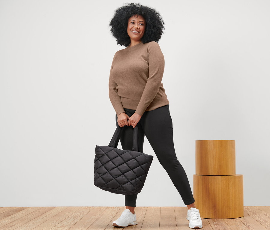 Une femme pose en pull en maille marron, legging noir et baskets blanches, tenant un sac fourre-tout noir matelassé.