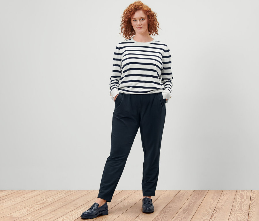 Une femme se tient debout, les mains dans les poches d'un pantalon de jogging, portant un pull en tricot fin à rayures, blanc.