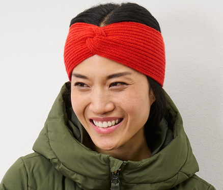 Une femme souriante, les bras croisés, portant un bandeau en maille orange et une doudoune verte.