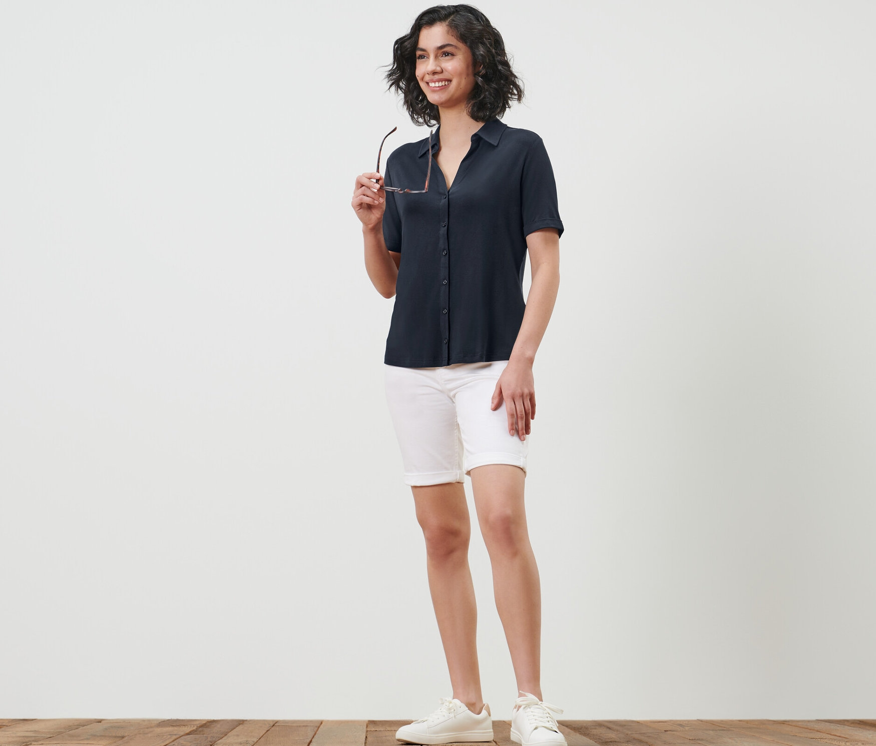 Femme tenant des lunettes et portant une blouse bleu foncé avec patte de boutonnage, un short blanc et des baskets.