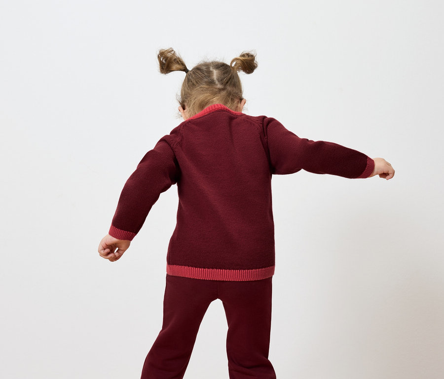 Une fille avec des couettes se tient dos à la caméra et agite les bras, vêtue d'un pull et d'un pantalon en tricot bordeaux.