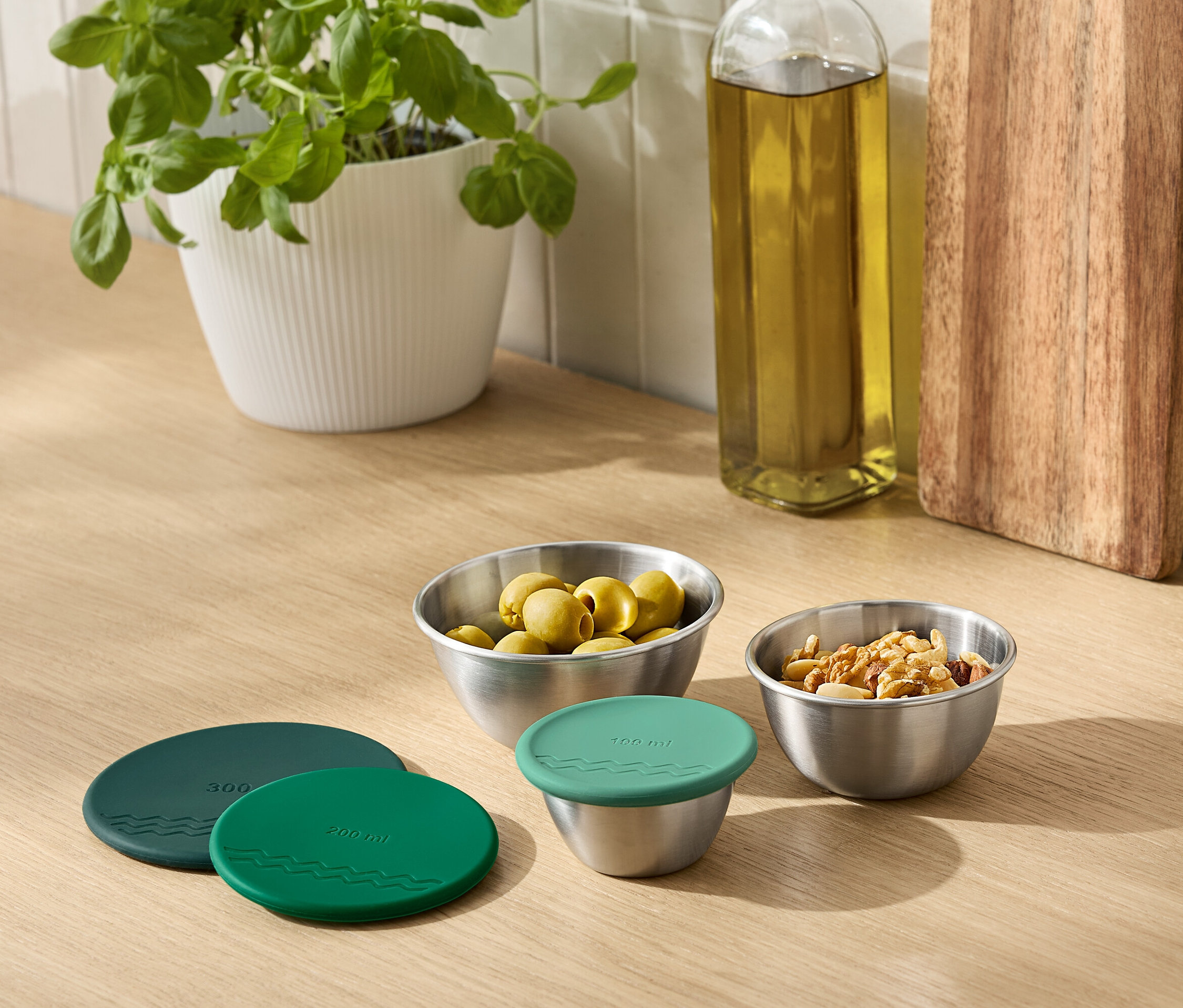 Sur un comptoir de cuisine se trouvent une plante en pot, une bouteille d'huile, une planche à découper et des bols en métal contenant des olives et des noix. Un bol est recouvert d'un couvercle vert.