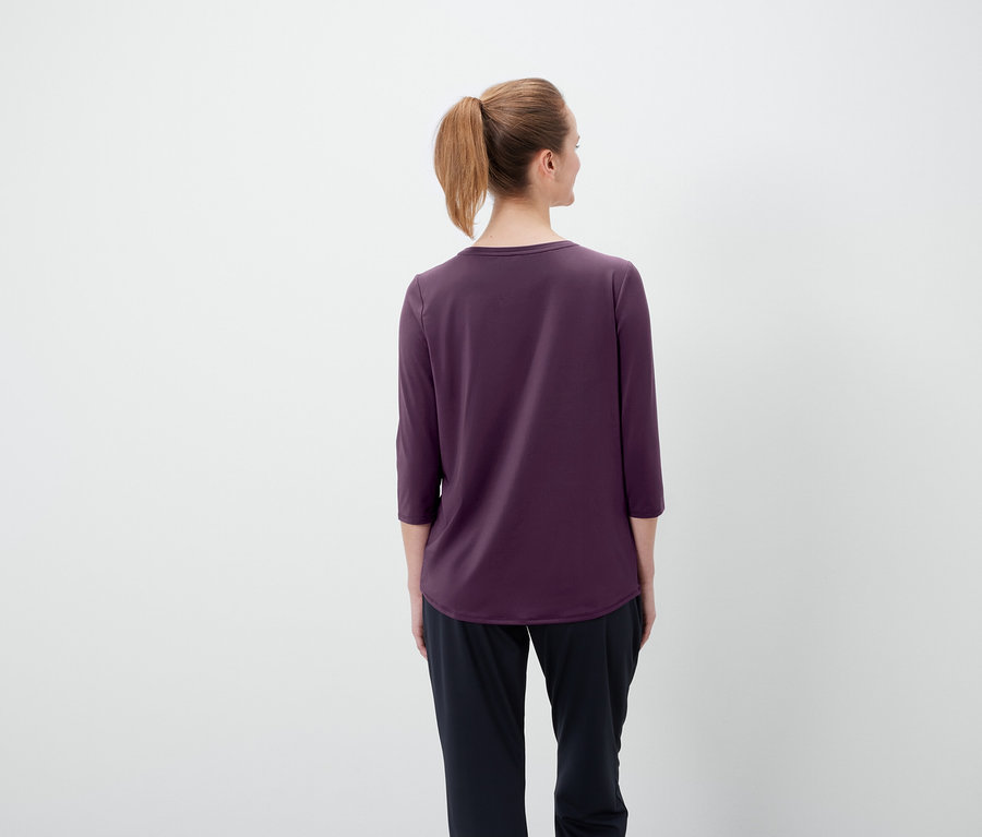 Vue de dos d'une femme aux cheveux bruns attachés en queue de cheval, portant un t-shirt violet à manches trois-quarts et un pantalon noir.