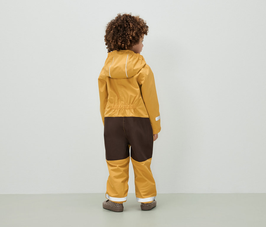 Un enfant aux cheveux bouclés se tient dos à la caméra et porte une combinaison marron et moutarde à capuche.