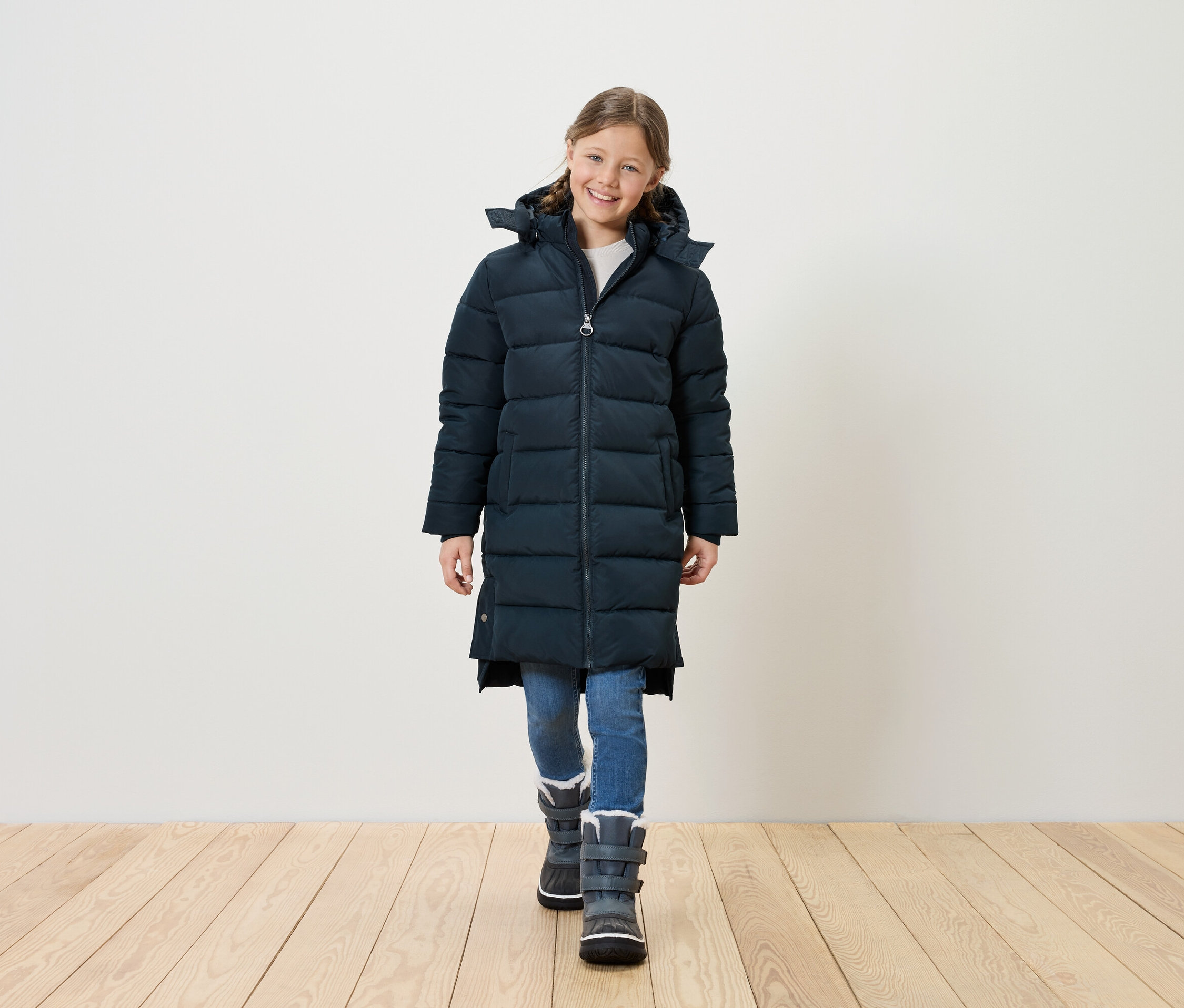 Une jeune fille souriante portant une longue doudoune bleu marine, un jean bleu et des bottes d'hiver grises se tient sur un plancher en bois devant un mur blanc.