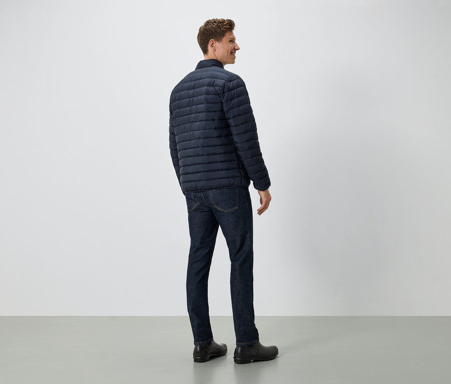 Un homme se tient dos à la caméra, portant une veste en duvet bleu marine, un jean et des chaussures noires.