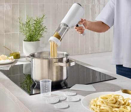 Une personne met des pâtes dans une casserole d'eau à l'aide d'une machine à pâtes. Une assiette de pâtes est posée sur le plan de travail.