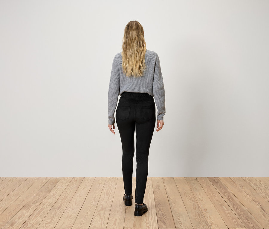 Une femme blonde vue de dos, portant un pull gris et un pantalon noir, marchant sur un plancher en bois.