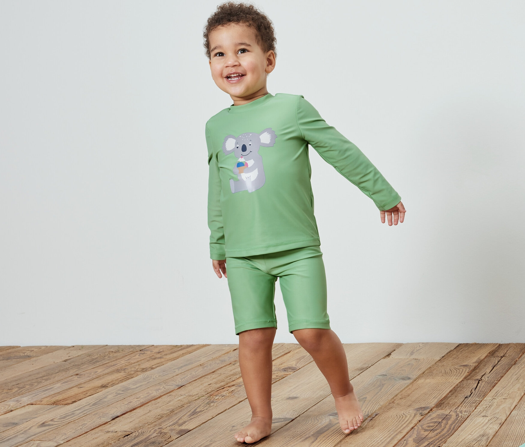 Un garçon portant une combinaison pour enfants avec protection UV 80, koala, se tient sur un plancher en bois à côté d'un jouet.