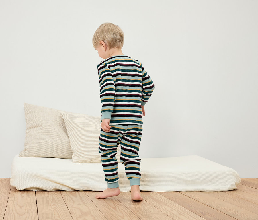 Un garçon se tient sur le sol en pyjama rayé à côté d'un lit blanc et d'oreillers.