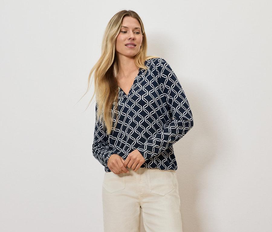 Une femme blonde pose en chemisier à motif géométrique et pantalon clair.