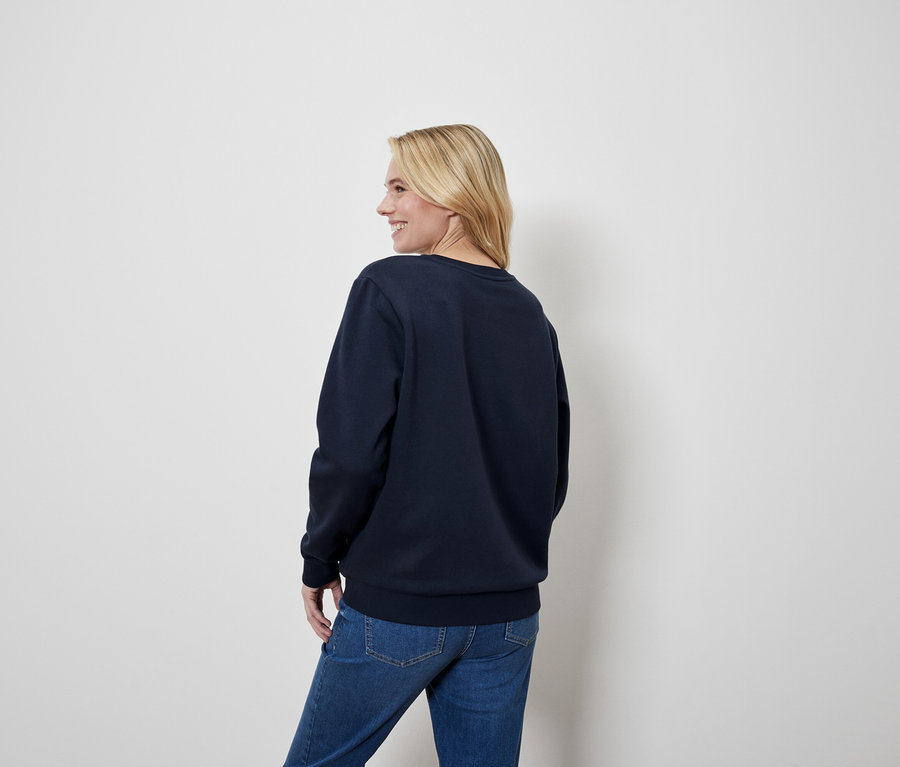 Une femme blonde regarde par-dessus son épaule, portant un pull bleu marine et un jean.