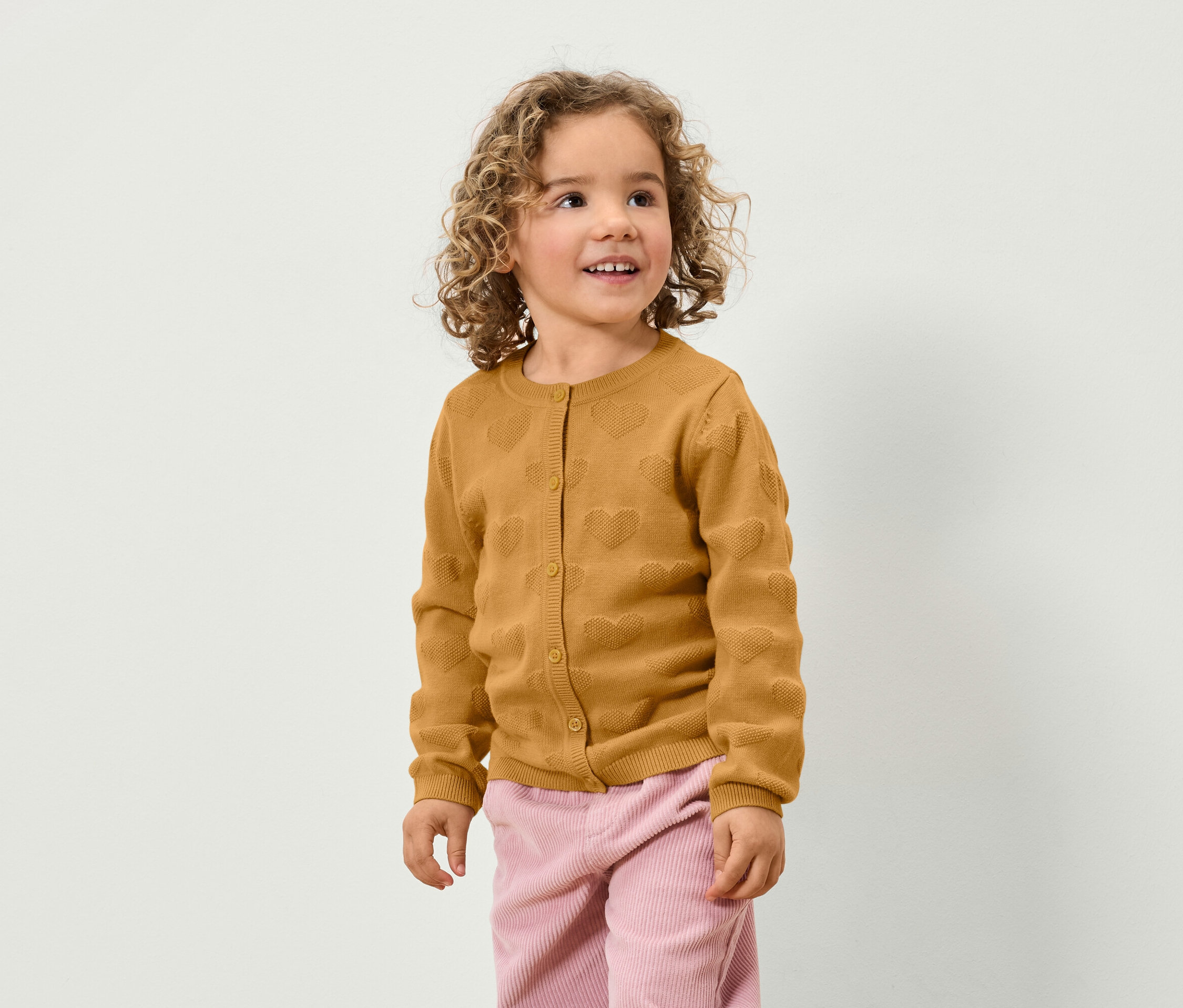 Une fillette aux cheveux bouclés porte un cardigan marron avec des coeurs et un pantalon rose.