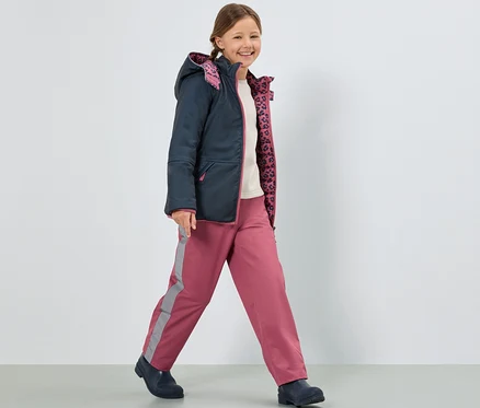 Une fille souriante marche et porte une veste bleu foncé et un pantalon de pluie rose pour enfants.