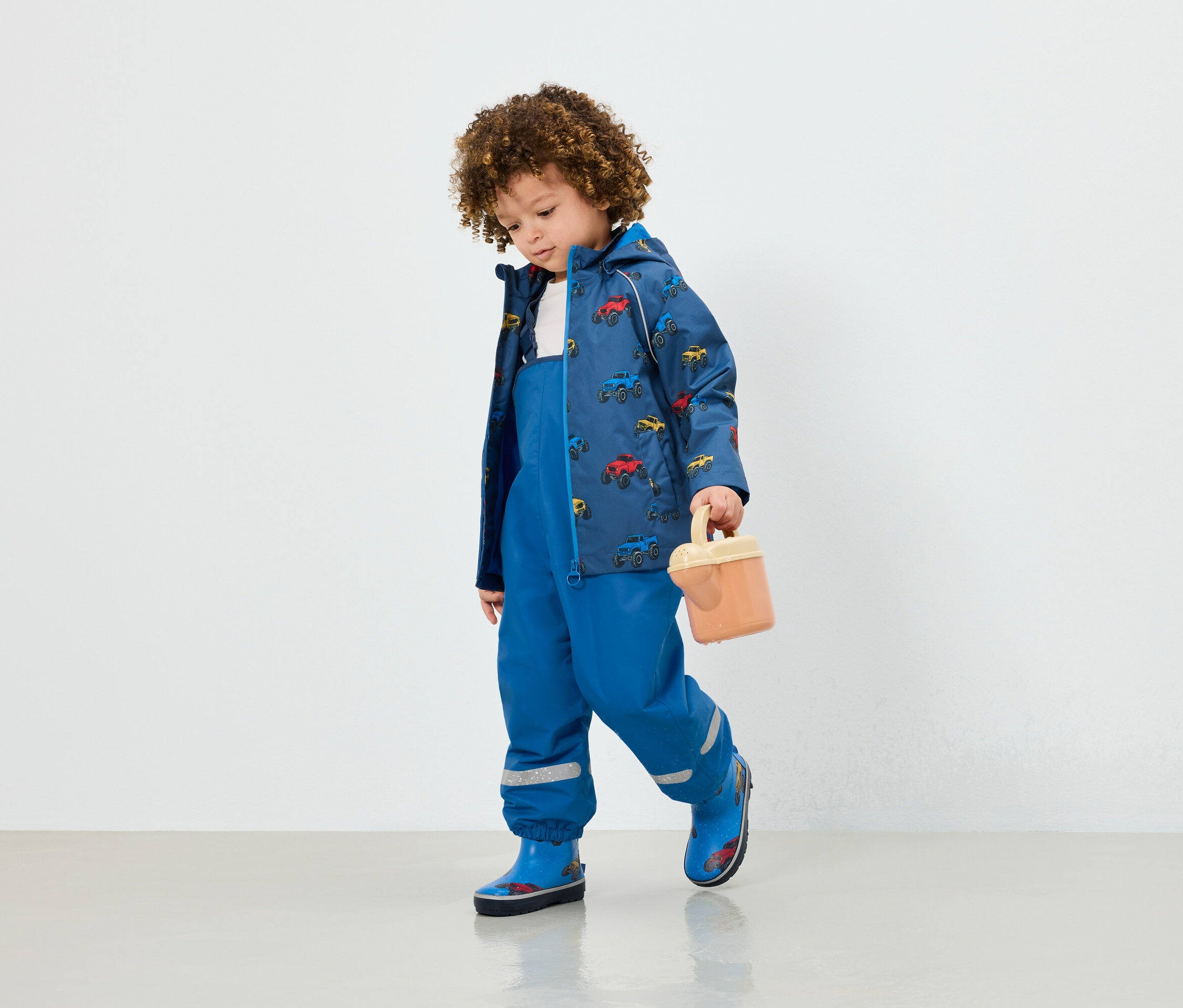 Un garçon en salopette bleue, une veste avec un imprimé de voiture et des bottes en caoutchouc bleues tient un jouet beige.