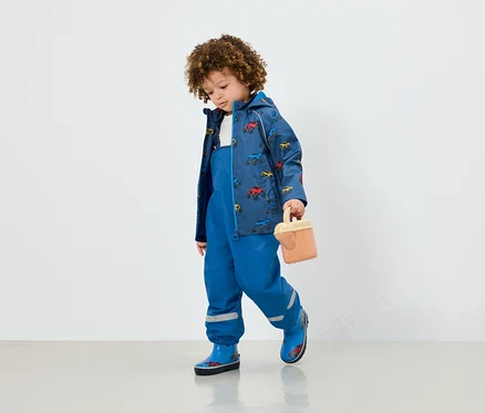 Un garçon en salopette bleue, une veste avec un imprimé de voiture et des bottes en caoutchouc bleues tient un jouet beige.