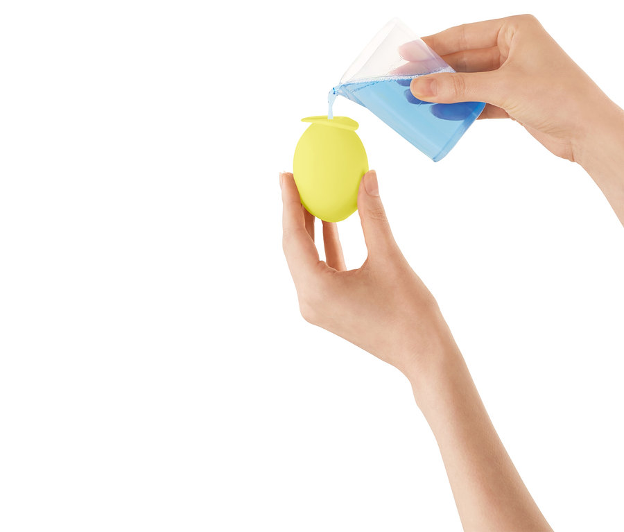 Des mains versent un liquide bleu d'un verre doseur dans un récipient jaune en forme d'œuf.