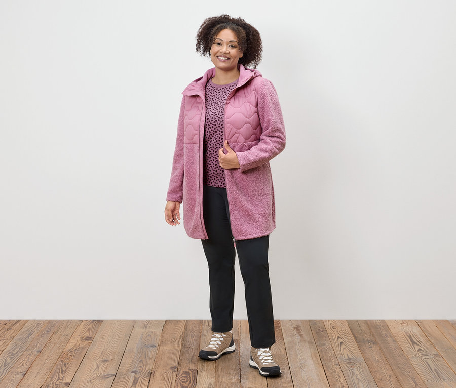 Une femme portant un manteau rose, un haut à motifs et un pantalon noir se tient sur un plancher en bois.