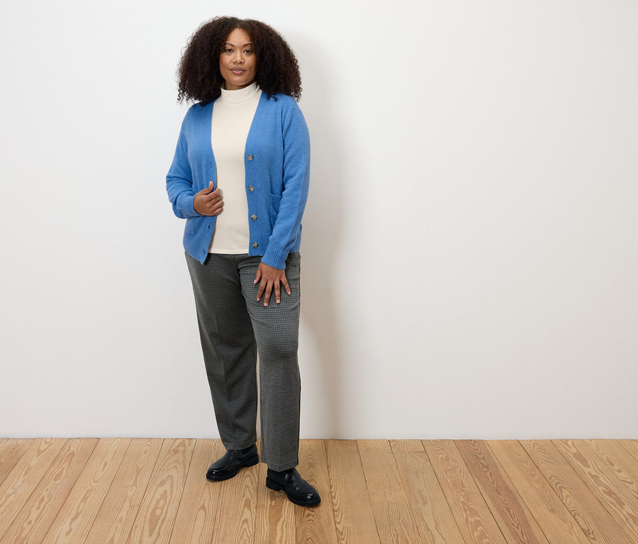 Une femme debout sur un plancher en bois porte un pantalon en jersey confortable à carreaux, un col roulé blanc et un cardigan bleu.