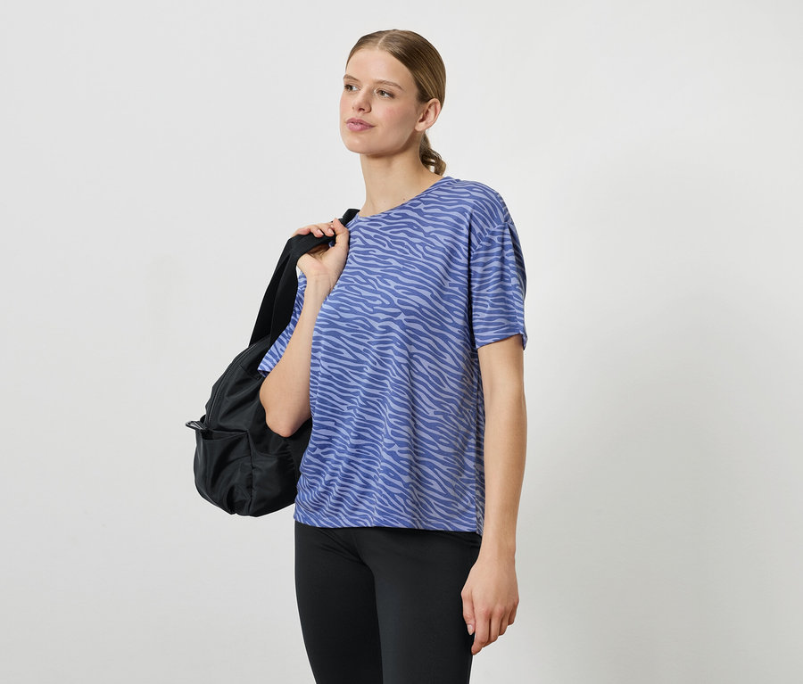 Une femme portant un t-shirt bleu à motifs et des leggings noirs, avec un sac à dos noir sur l'épaule.