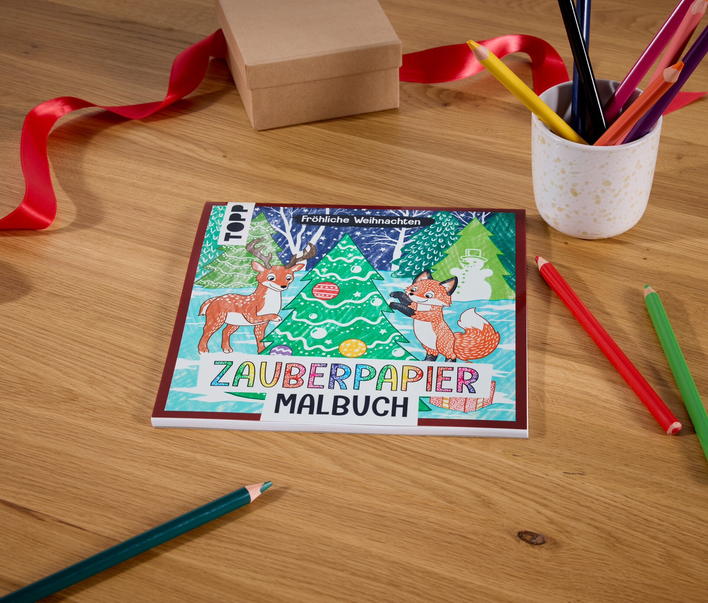 Sur une table en bois se trouvent un livre de coloriage Zauberpapier Malbuch avec un motif de Noël, une boîte avec un ruban rouge, un gobelet avec des crayons de couleur et deux crayons de couleur.