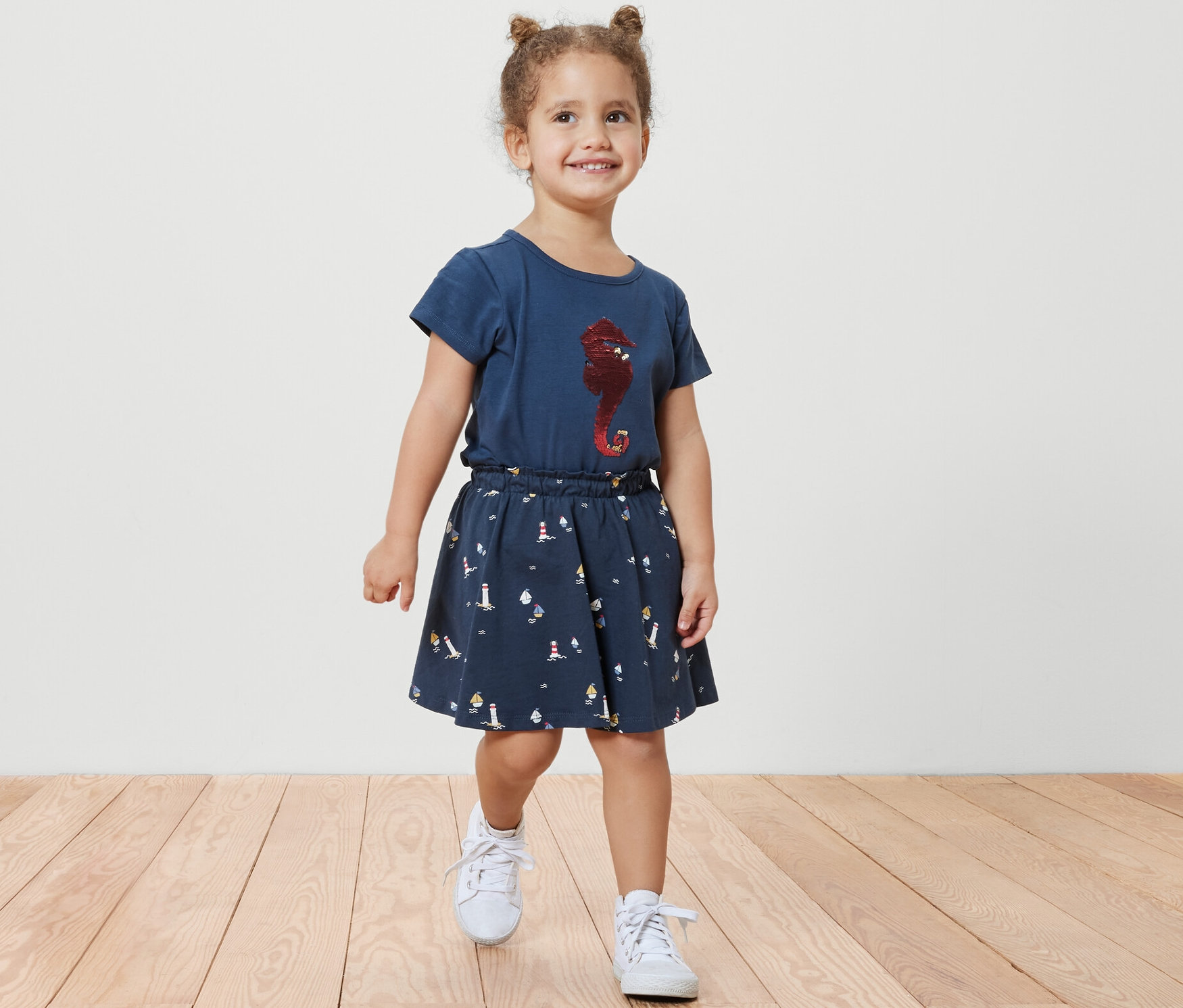 Une petite fille souriante marche sur un plancher en bois, portant une jupe en jersey pour enfants avec pantalon, maritime, et un t-shirt bleu avec un imprimé d'hippocampe.