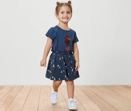 Une petite fille souriante marche sur un plancher en bois, portant une jupe en jersey pour enfants avec pantalon, maritime, et un t-shirt bleu avec un imprimé d'hippocampe.