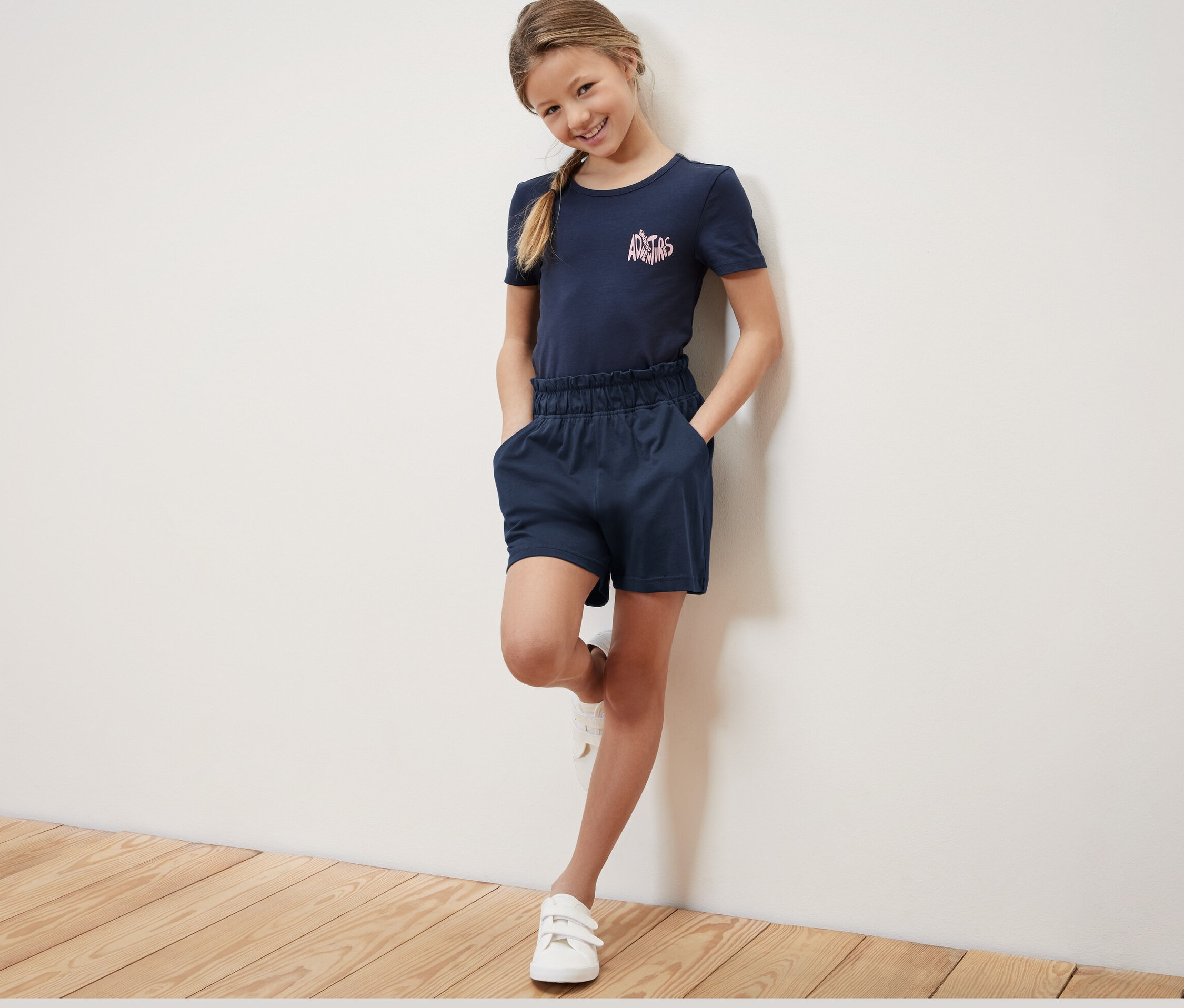 Une fille souriante pose dans un t-shirt bleu marine et 2 shorts en jersey pour enfant. Elle a les mains dans les poches du short.