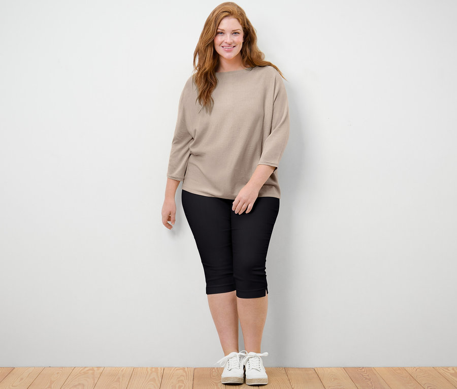 Une femme debout portant un pull en maille fine beige chiné et un pantalon 3/4 stretch noir.