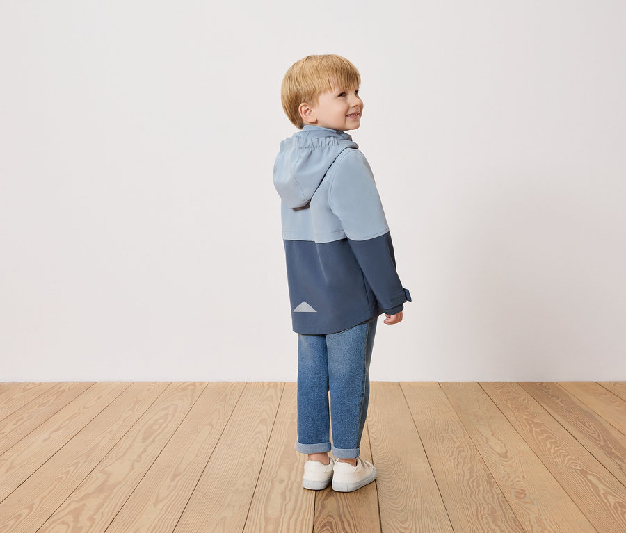 Un garçon souriant se tient de profil, portant une veste bleue bicolore, un jean et des chaussures blanches.