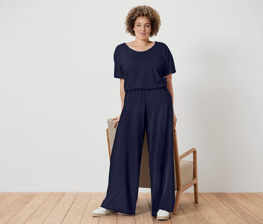 Une femme aux cheveux bouclés porte une combinaison bleu marine et des chaussures blanches, elle se tient à côté d'une chaise au cadre en bois.