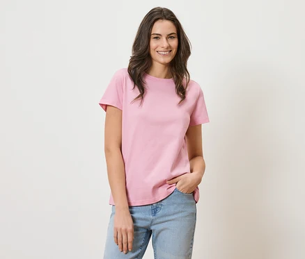 Femme aux longs cheveux bruns portant un t-shirt rose et un jean bleu souriant.