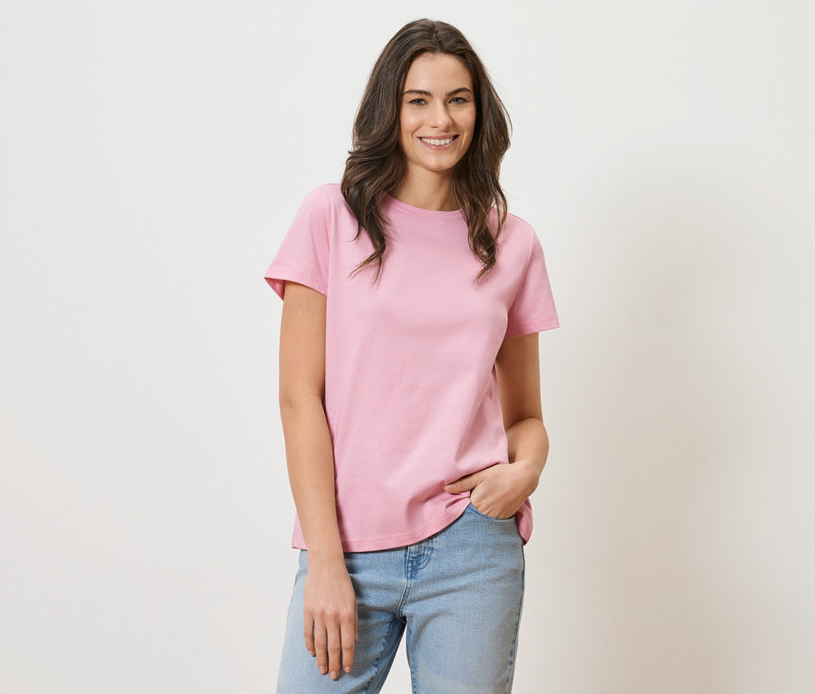 Femme aux longs cheveux bruns portant un t-shirt rose et un jean bleu souriant.