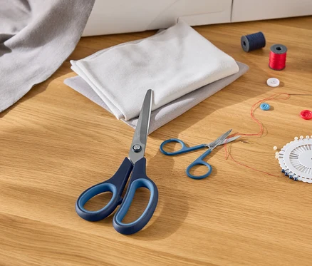 Sur une table en bois sont posés des morceaux de tissu, de grands ciseaux à manche bleu, de petits ciseaux, des aiguilles, des fils, des boutons et des bobines de fil.