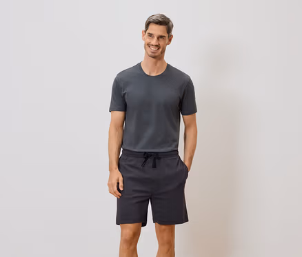 Un homme souriant portant un t-shirt gris et un short à cordon se tient devant un mur blanc.