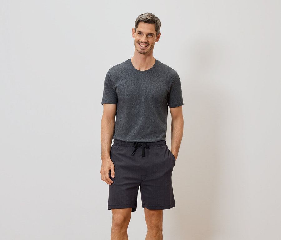 Un homme souriant portant un t-shirt gris et un short à cordon se tient devant un mur blanc.