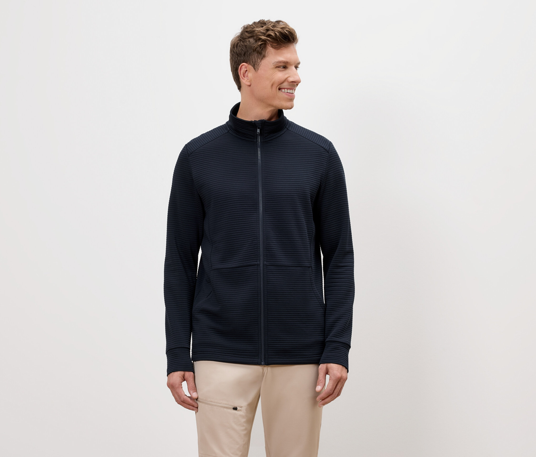 Un homme aux cheveux blonds porte une veste zippée bleu marine et un pantalon beige.